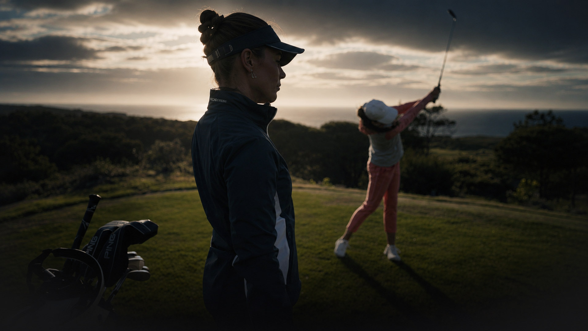 Two golfers on a coastal golf course at sunset, one watching while the other swings a club in the background with a cart nearby.