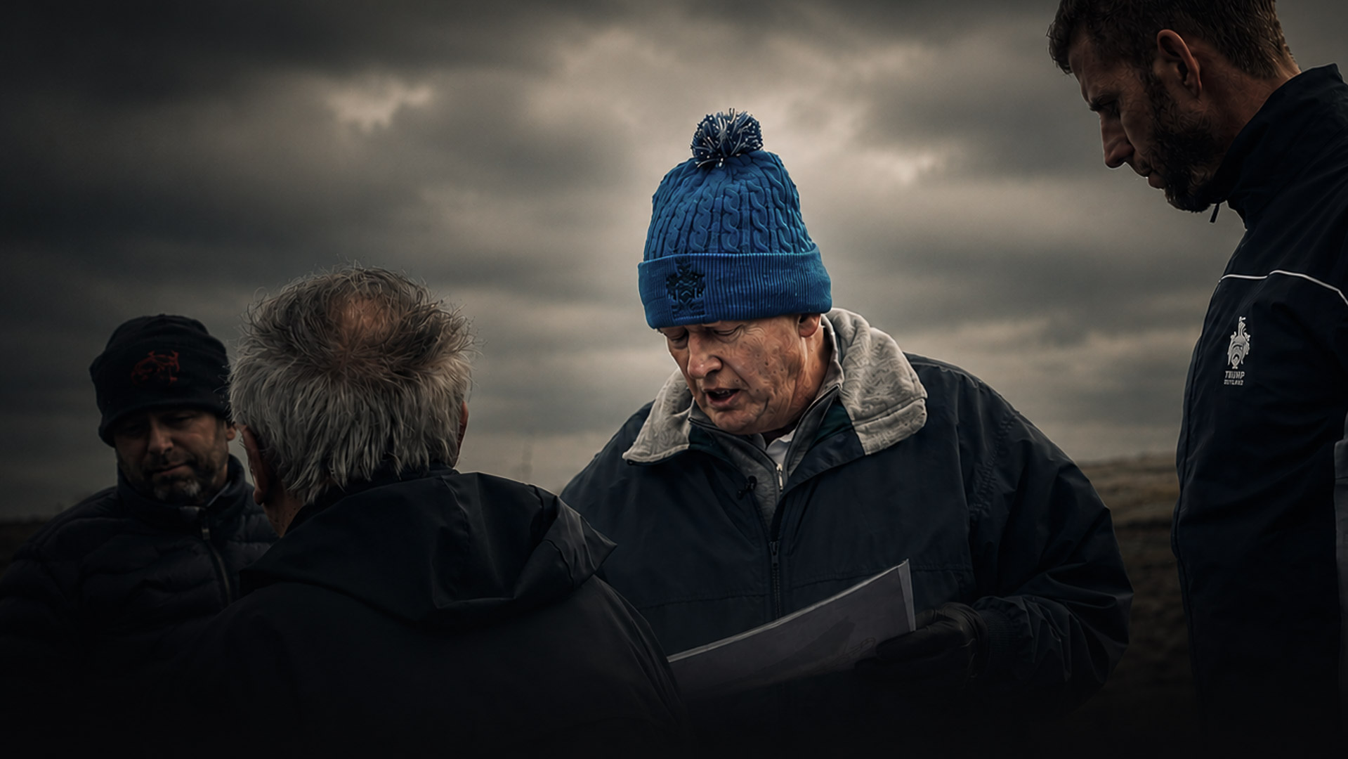 Group of four outdoors on a dark, cloudy day; man in a blue knit hat reads a map or document while others listen.